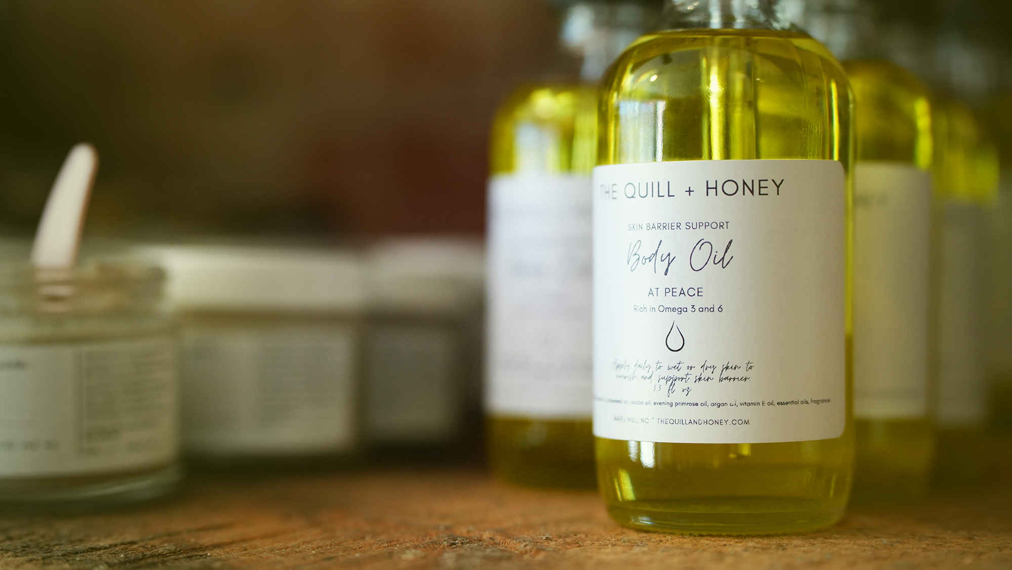 Clear glass bottles filled with yellow body oil are arranged on a wooden surface. Captured with expert photography & videography services, the front bottle is labeled “Quill + Honey Body Oil At Peace,” with blurred jars and more bottles in the background.