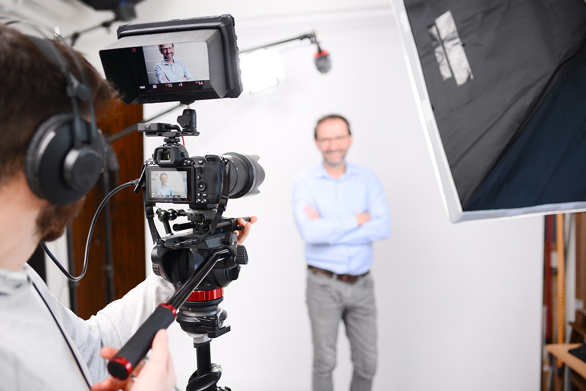 A person with headphones films a man standing with arms crossed in a studio setup, using a camera on a tripod. The man appears on the camera screen, and bright lighting equipment surrounds the scene.